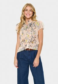 Smiling woman with long blonde hair wears a ruffled, floral short-sleeve blouse tucked into high-waisted dark blue wide-leg pants.
