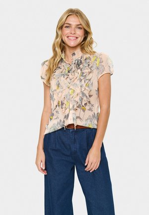 Lächelnde Frau mit langen blonden Haaren trägt eine plissierte, floral gemusterte Bluse mit kurzen Ärmeln, die in eine hoch taillierte dunkelblaue weite Hose gesteckt ist.