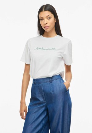 Kvinne med langt mørkt hår iført en hvit T-skjorte med teksten "Mademoiselle" og blå vidbukser med høy midje, står med en hånd i lommen.