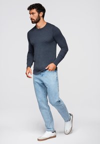 Camicia a maniche lunghe blu scuro abbinata a jeans blu chiaro e sneaker bianche con dettagli marroni, indossata da un uomo che cammina.