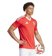 Rotes Fußballtrikot mit weißen Akzenten und V-Ausschnitt, ausgestattet mit einem strukturierten Zickzackmuster, kurzen Ärmeln und einem Vereinslogo auf der Brust.
