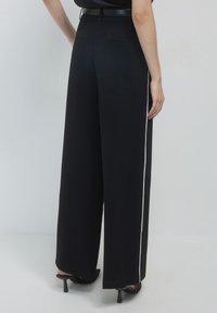 Persona con pantaloni larghi neri con sottili strisce bianche laterali, una cintura nera e scarpe con tacco nere, in piedi su uno sfondo uniforme.