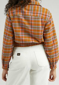 Cropped plaid shirt in orange, gray, and pink with button closure and long sleeves. Paired with white jeans featuring back pockets and logo patch.