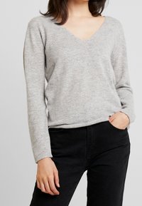 Hellgrauer V-Ausschnitt-Pullover aus weichem Strickstoff, mit langen Ärmeln und lässiger Passform. Kombiniert mit schwarzen Hosen.