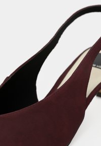 Κοντινή λήψη ενός μπορντό σουέτ γυναικείου παπουτσιού slingback με χαμηλό τακούνι και μαύρη εσωτερική επένδυση σε λευκό φόντο.