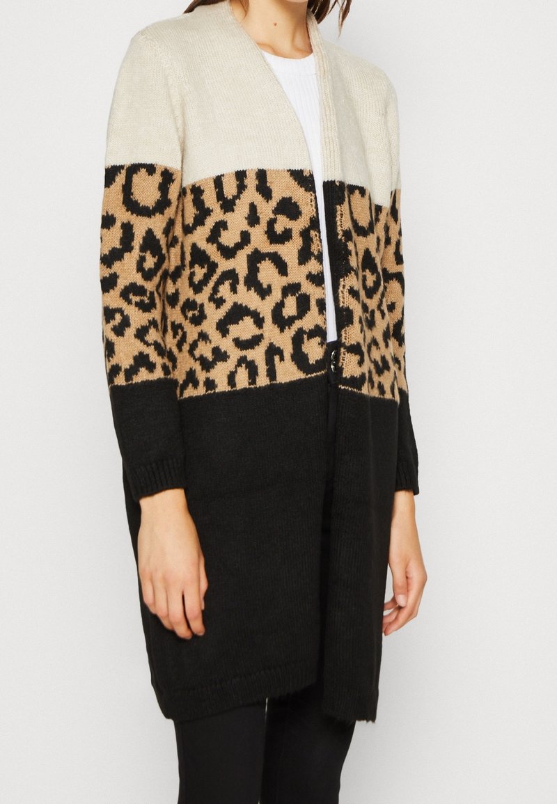 Femme portant un long cardigan ouvert à l'avant avec des blocs de couleur crème, imprimé léopard et noir, sur un haut blanc et un pantalon noir.
