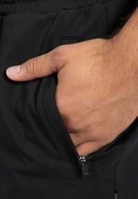 Zwarte short met een zijzak, met een ritssluiting. De stof is glad met een subtiele glans, en een hand steekt in de zak.