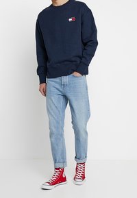 Marinblå sweatshirt med en liten logotyp, ljusblå jeans och röda högskaftade sneakers. Avslappnad passform, bomullsmaterial och upplagda ärmar.