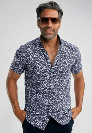 Homme d'âge moyen portant des lunettes de soleil et une chemise bleu marine à manches courtes avec un motif floral blanc, les mains posées négligemment dans les poches.