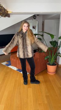 Faux fur coat in brown shades, featuring a relaxed fit and wide sleeves. Paired with black velvet pants and black shoes, on a wooden floor.