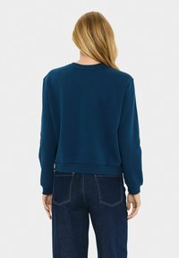 Donna con lunghi capelli biondi, che indossa una semplice felpa blu scuro e jeans blu scuro, vista da dietro su uno sfondo bianco.