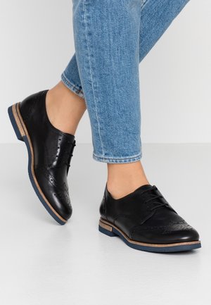 Personne portant des chaussures oxford brogue en cuir noir avec une semelle bleue, associées à un jean en denim bleu clair, sur fond blanc.