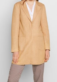 Abrigo de mezcla de lana beige con un corte entallado, con solapas de muesca, dos bolsillos frontales y una textura suave, llevado sobre pantalones a cuadros.