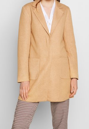 Cappotto in misto lana beige con vestibilità su misura, rever a lancia, due tasche anteriori e una texture liscia, indossato sopra pantaloni a quadri.