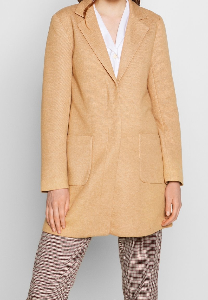 Abrigo de mezcla de lana beige con un corte entallado, con solapas de muesca, dos bolsillos frontales y una textura suave, llevado sobre pantalones a cuadros.
