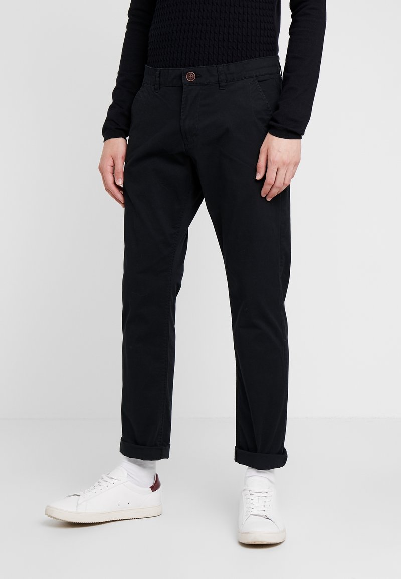 Personne portant un pantalon noir avec revers, des chaussettes blanches, des baskets blanches avec un détail bordeaux et un haut noir à manches longues sur un fond uni.