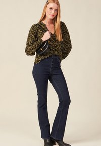 Borsa nera, blusa verde con motivo e maniche elastiche, e jeans a zampa d'elefante in denim scuro con bottoni frontali. Modello con stivali neri alla caviglia.