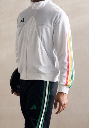 Biała sportowa kurtka z szarymi akcentami i kolorowymi paskami na rękawach. W połączeniu z czarnymi spodniami z zielonymi paskami i logo.