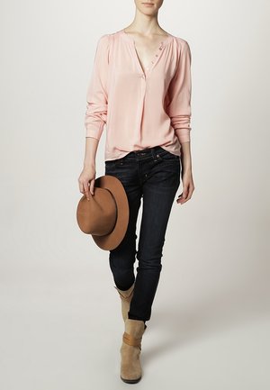 Blouse rose clair à manches longues avec un décolleté en V et des détails de boutons, assortie à un jean foncé et des bottines beiges, tenant un chapeau brun.