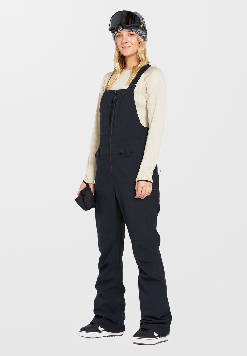 Navy wasserfeste Schnee-Overalls mit Reißverschluss, verstellbaren Trägern und mehreren Taschen, kombiniert mit einem leichten Langarmshirt und einer Schutzbrille.