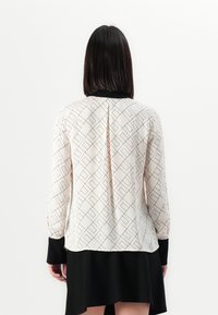 Long-sleeve blouse featuring a geometric pattern in cream with black accents, collar, and a slit at the back, worn with a black skirt.