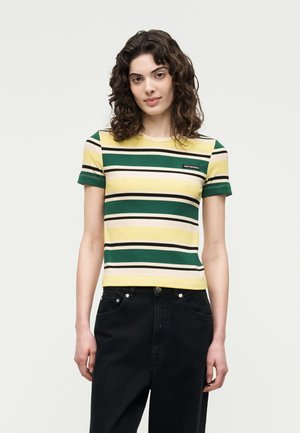 Mujer con cabello rizado que lleva una camiseta de manga corta y acanalada con rayas horizontales en verde, amarillo, negro y blanco, y pantalones negros.