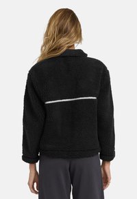 Schwarze Fleecejacke mit strukturiertem Material, ausgestattet mit einem weißen horizontalen Streifen auf dem Rücken und einer entspannten, boxy Passform.