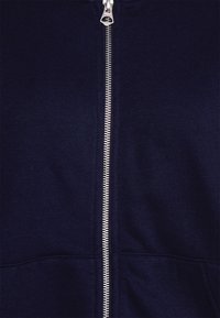 Marineblå lynlås-hættetrøje lavet af blødt stof, med en sølvfarvet lynlås og kængurulommer foran. Glat tekstur med et afslappet design.