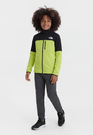 Lokkis juustega laps, kellel on seljas must ja laimirohelise lukuga jope, tumedad hallid püksid ja mustad tossud, seisab ja naeratab.