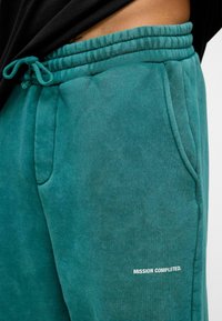 Teal joggingbroek met elastische tailleband en trekkoord. Voorzien van zijzakken en bedrukte tekst "MISSIE VOLTOOID." in het wit. Soepele textuur.