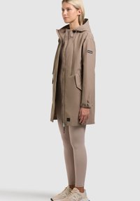 Žena stojící v béžovém plášti s kapucí a předními kapsami, doplněném světle béžovými legíny a béžovými teniskami na neutrálním pozadí.