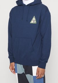 Sudadera con capucha azul marino hecha de algodón suave, con un parche gráfico triangular en verde lima y negro, con un bolsillo delantero y capucha con cordón.