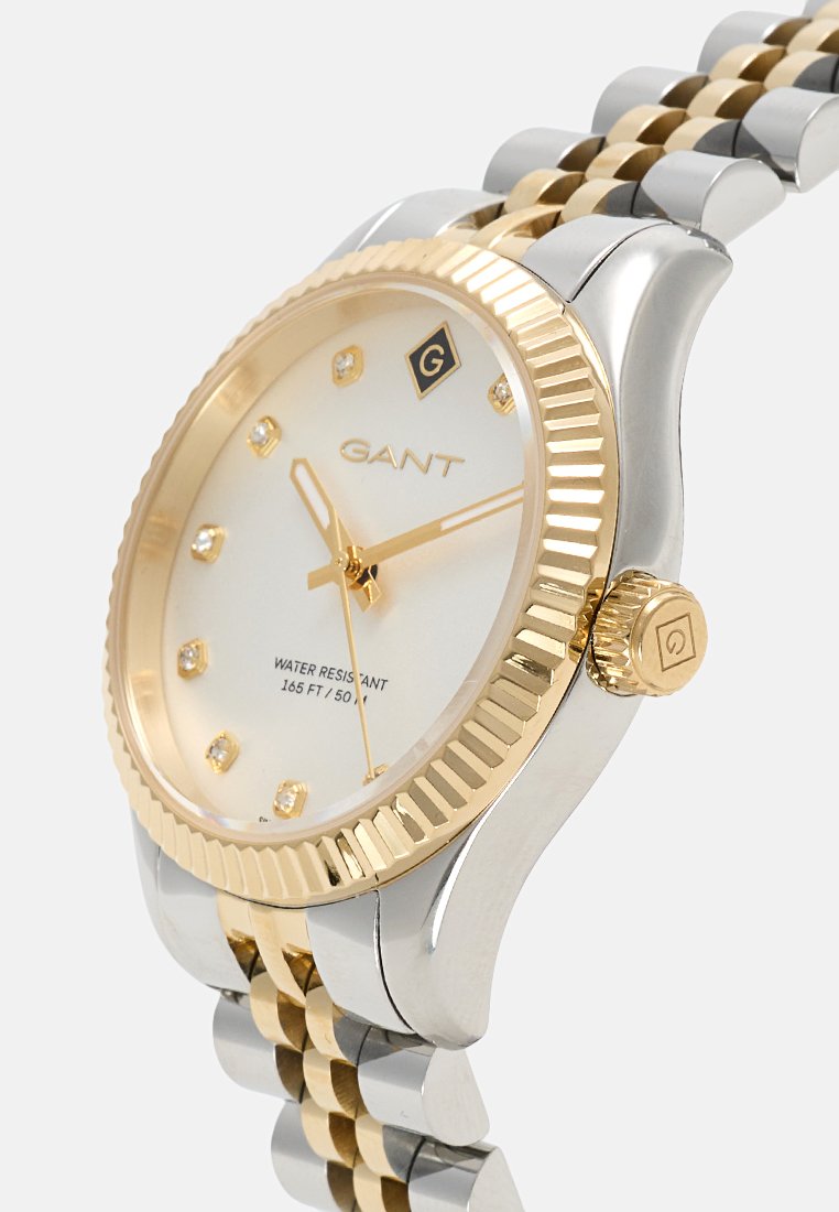 Montre en or et argent avec un cadran blanc, des marqueurs semblables à des diamants et une lunette texturée en or. Dispose d'un bracelet en métal bi-ton. Résistante à l'eau.