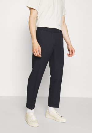 Man draagt zwarte, op maat gemaakte broek met omslag bij de zoom, witte sokken, crèmekleurige sneakers en een wit overhemd met korte mouwen, ingestopt, tegen een effen achtergrond.