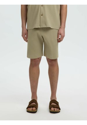 Homme portant un short côtelé beige et une chemise assortie avec des sandales ouvertes marron, debout sur fond blanc.