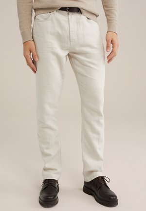 Mann trägt beige Hose, beigen Pullover, schwarzen Gürtel und schwarze Schnürschuhe und steht vor einem schlichten Hintergrund.