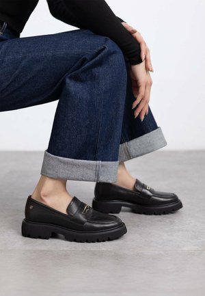 Mocassins en cuir noir avec un profil bas et une semelle en caoutchouc épaisse. Présente un design élégant et un accent doré sur la sangle.