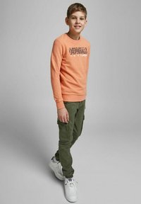 Maglione arancione a girocollo con testo stampato, abbinato a pantaloni cargo verde oliva. Sneakers bianche completano l'outfit.