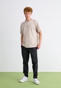 Beige bombažna majica s zelenim logotipom "Converse", v kombinaciji s črnimi hlačami in belimi športnimi copati. Model stoji na zeleni tla s preprosto ozadje.