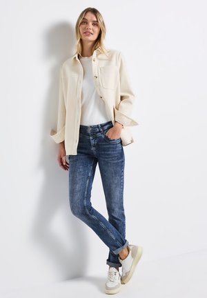 Veste beige clair avec deux poches avant, t-shirt blanc et jean skinny bleu. Des baskets décontractées avec des accents crème et gris complètent la tenue.