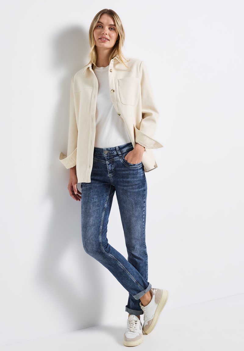 Giacca beige chiaro con due tasche frontali, t-shirt bianca e jeans skinny blu. Sneakers casual con accenti crema e grigio completano l'outfit.