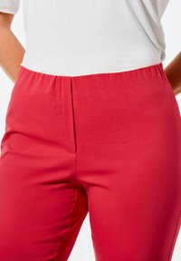 Rode stretchbroek met een elastische tailleband; gladde textuur; vlakke voorkant met een naaddetail langs de zijkant. Gecombineerd met een zichtbaar wit shirt.