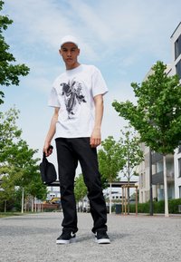 Hvid t-shirt med et sort grafisk design, sorte jeans, sorte sneakers med hvide detaljer, og en sort kasket i venstre hånd.