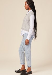 Maglione grigio a maglia senza maniche sopra una camicia bianca a righe, abbinato a jeans a gamba affusolata di un blu chiaro e scarpe slip-on nere. Capelli lunghi intrecciati.