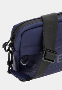 Bolso cruzado sintético en color navy con superficie texturizada, correa negra ajustable, cierre de cremallera y detalle de logo en relieve.