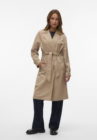 Trench coat beige con vita regolabile, due tasche frontali e colletto a revers, abbinato a jeans scuri e scarpe nere.