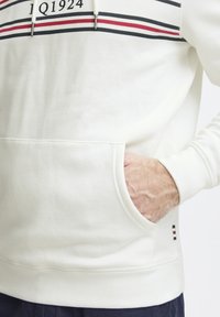 Sudadera de algodón blanca con un bolsillo frontal, que presenta rayas horizontales en rojo y negro en el pecho y un logo encima.