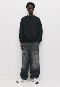 Schwarzer Pullover mit Rundhalsausschnitt, graue Cargo-Jeans mit großen Taschen und weiße Sneaker. Der Stoff wirkt weich und strapazierfähig.