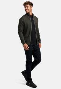 Donkergroene textuurjas met een hoge kraag en zilveren ritssluiting, gedragen over een zwart shirt en gecombineerd met donkere broek en zwarte sneakers.