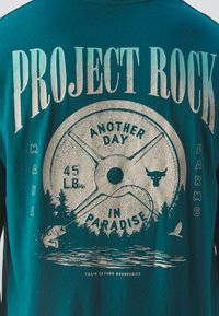 Tyrkysové tričko s krátkym rukávom, na ktorom je grafický dizajn s váhami, rybou, stromami a horami. Text znie "PROJECT ROCK" a "ANOTHER DAY IN PARADISE."
